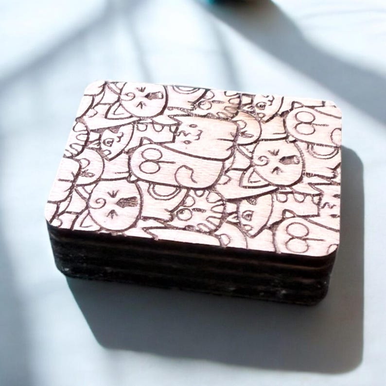 May include: A stack of wooden coasters with a laser-etched design of cartoon cats. The coasters are square and have a light brown color with dark brown etched details. The design features various cat faces in a repeating pattern.