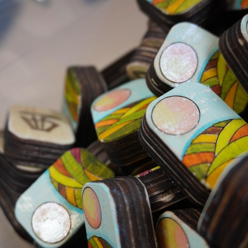Op de afbeelding: Een verzameling kleine, rechthoekige houten objecten met kleurrijke, handgeschilderde ontwerpen. Elk stuk heeft een lichtblauwe achtergrond met een cirkelvormig ontwerp en een landschapspatroon in geel, oranje en groen.