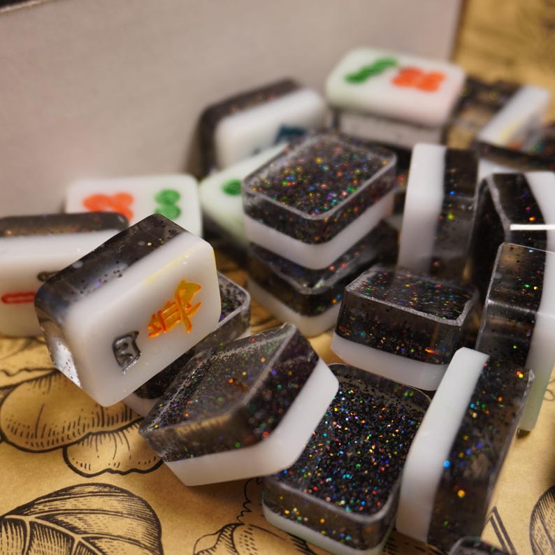Pu&ograve; includere: Una collezione di tessere Mahjong colorate. Le tessere presentano un mix di resina bianca, nera e glitterata, con vari simboli e caratteri in arancione, verde e rosso. Le tessere sono disposte su una superficie a motivi.