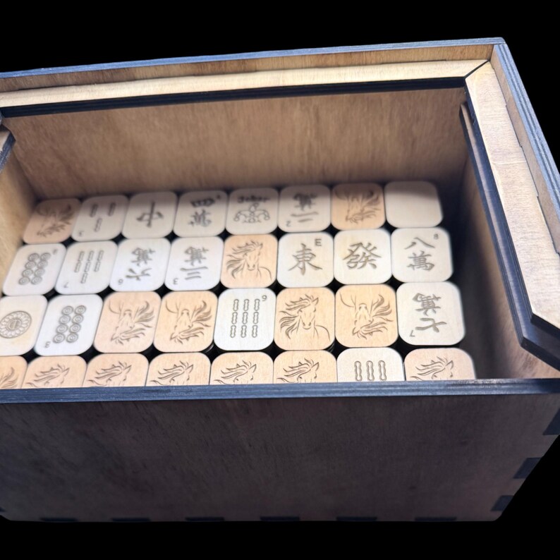 May include: A wooden box with a partially open lid, filled with light-colored Mahjong tiles. The tiles feature various symbols and characters, including horses and Chinese numerals. The box is made of light-colored wood.