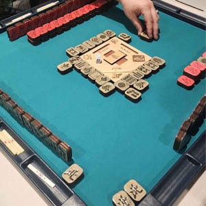May include: A Mahjong game in progress, featuring a teal playing surface. The game includes wooden tiles with Chinese characters, red tiles, and a wooden center board. A hand is reaching for a tile.