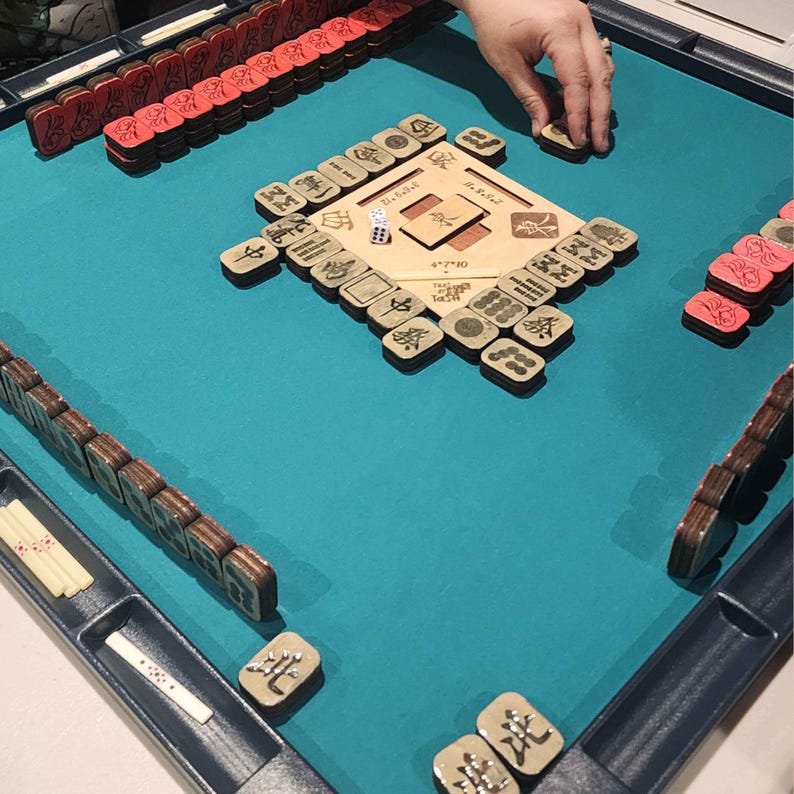 May include: A Mahjong game in progress, featuring a teal playing surface. The game includes wooden tiles with Chinese characters, red tiles, and a wooden center board. A hand is reaching for a tile.