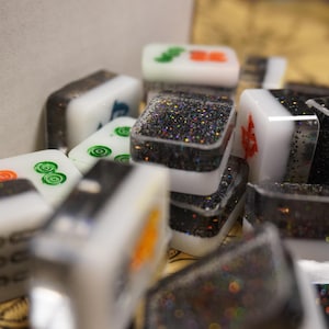 May include: Close-up of a collection of Mahjong tiles. The tiles are white with various symbols in green, orange, and blue. Some tiles have a black glitter design. The tiles are stacked and scattered, creating a textured appearance.