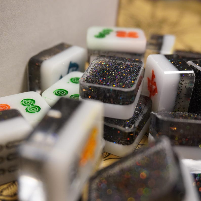 May include: Close-up of a collection of Mahjong tiles. The tiles are white with various symbols in green, orange, and blue. Some tiles have a black glitter design. The tiles are stacked and scattered, creating a textured appearance.
