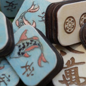May include: Close-up of wooden game tiles. Some tiles are light blue with a koi fish design and Asian characters. Other tiles are natural wood with etched floral and character designs. The tiles have dark brown edges.
