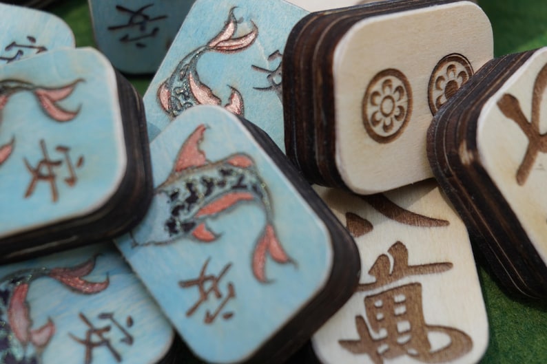 May include: Close-up of wooden game tiles. Some tiles are light blue with a koi fish design and Asian characters. Other tiles are natural wood with etched floral and character designs. The tiles have dark brown edges.