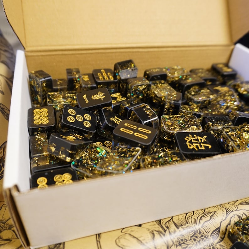 Op de afbeelding: Een doos gevuld met zwarte en gouden Mahjong-tegels. De tegels hebben gouden symbolen en cijfers. De tegels zijn rechthoekig en hebben een heldere hars met gouden glitter. De doos is van karton.