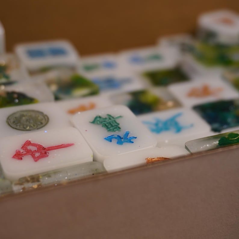 May include: Close-up of a set of white Mahjong tiles with colorful symbols and characters. The tiles have red, green, and blue accents. The game pieces are arranged on a surface with a blurred background.