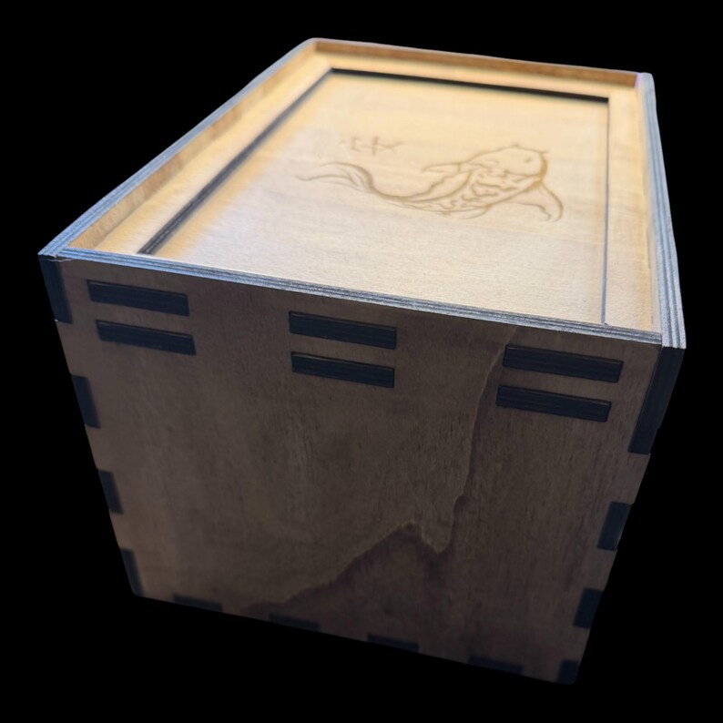 May include: Wooden box with a lid featuring a laser-etched koi fish design. The box is constructed from light-colored wood with dark accents at the corners. The lid is flush with the box, and the design is centered.