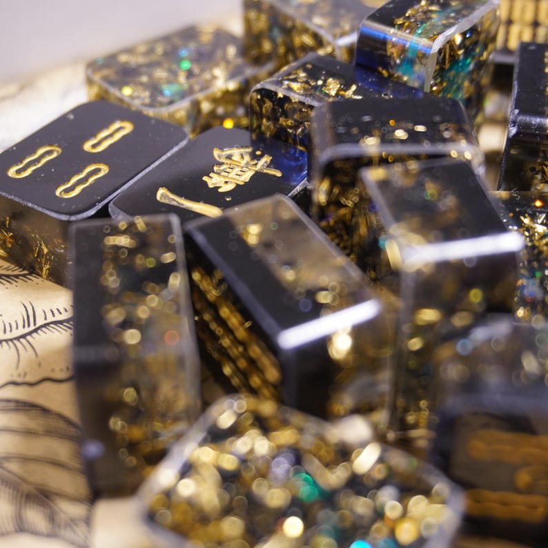 Op de afbeelding: Een verzameling zwarte dominostenen met gouden accenten en ingebedde gouden vlokken. Sommige dominostenen hebben gouden symbolen. De dominostenen zijn gemaakt van een helder harsmateriaal, wat een unieke en decoratieve uitstraling cre&euml;ert. Close-up.