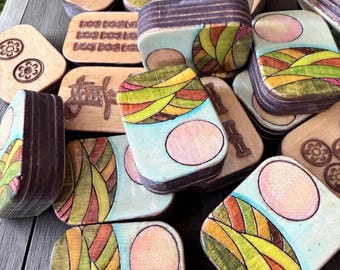 Puede incluir: Una colección de fichas rectangulares de madera con diversos diseños. Algunas fichas muestran paisajes pintados con colinas y soles circulares en tonos verde, amarillo y rosa. Otras presentan patrones grabados al fuego, incluyendo motivos florales y texto.