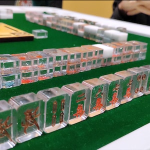 May include: Clear acrylic Mahjong tiles with red and brown symbols and characters are arranged on a green felt surface. The tiles are stacked in rows, showcasing the game's components.