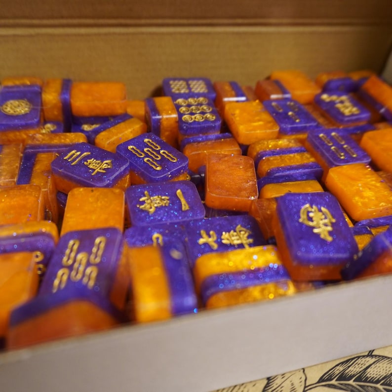 May include: A collection of colorful Mahjong tiles in a wooden box. The tiles are a mix of orange and purple with gold-colored symbols. The tiles are arranged in a random pattern, suggesting a game in progress.