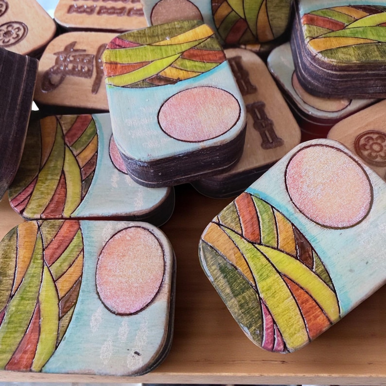 Handmade wooden Riichi Mahjong tiles featuring a colorful sunrise design with pink sun and rolling hills.