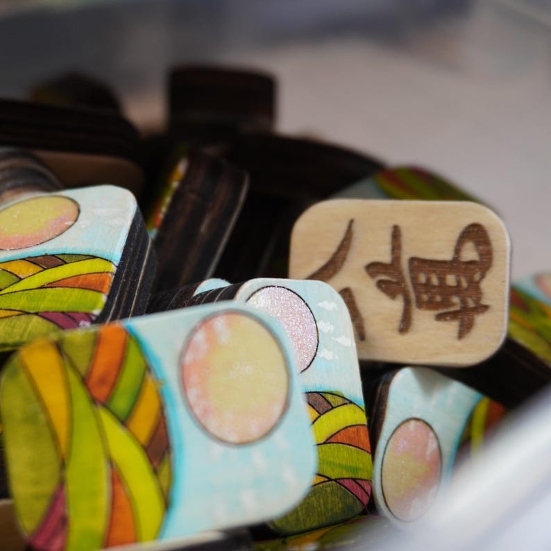 Op de afbeelding: Een verzameling kleine houten spelstukken. Sommige stukken tonen een landschapsontwerp met een gele zon, groene en oranje heuvels en een blauwe lucht. E&eacute;n stuk heeft Japanse karakters.