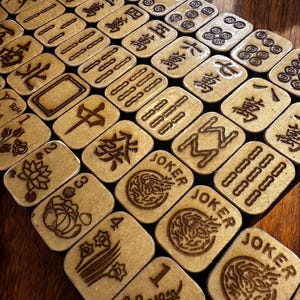 May include: A collection of wooden Mahjong tiles with engraved symbols and characters. The tiles are beige with dark brown etched designs, including numbers, suits, and the word "JOKER". The tiles are arranged on a dark wooden surface.
