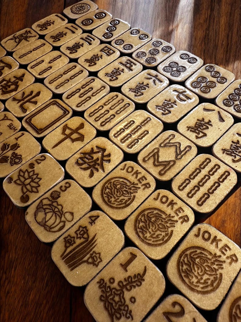 May include: A collection of wooden Mahjong tiles with engraved symbols and characters. The tiles are beige with dark brown etched designs, including numbers, suits, and the word "JOKER". The tiles are arranged on a dark wooden surface.