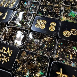 Op de afbeelding: Close-up van meerdere zwarte rechthoekige hars tegels met gouden accenten en ingebed bladgoud en glitter. Sommige tegels hebben gouden symbolen of patronen. De tegels zijn dicht bij elkaar geplaatst, waardoor een moza&iuml;ekachtig effect ontstaat.