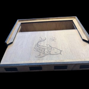 May include: Wooden box with a laser-etched koi fish design and Japanese characters. The box is made of light-colored wood with dark edges and a rectangular shape. The lid is partially open, revealing the interior.