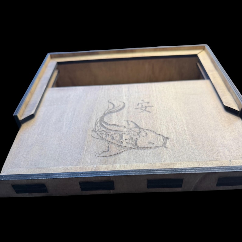 May include: Wooden box with a laser-etched koi fish design and Japanese characters. The box is made of light-colored wood with dark edges and a rectangular shape. The lid is partially open, revealing the interior.
