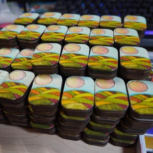 Op de afbeelding: Een verzameling rechthoekige houten dozen, elk met een landschapsschilderij. Het kunstwerk toont glooiende groene en gele heuvels onder een lichtblauwe lucht met een zon. De dozen hebben donkerbruine zijkanten en zijn opgestapeld.