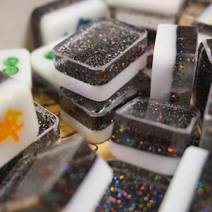 May include: Close-up of several Mahjong tiles. The tiles are rectangular with a clear resin exterior filled with black glitter. Each tile has a white band in the middle. Some tiles have green or orange symbols.