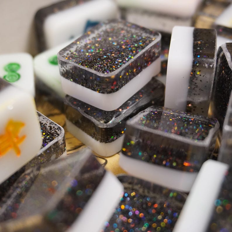 May include: Close-up of several Mahjong tiles. The tiles are rectangular with a clear resin exterior filled with black glitter. Each tile has a white band in the middle. Some tiles have green or orange symbols.