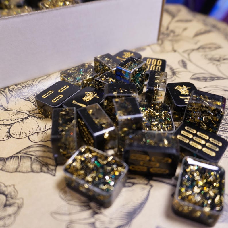 Op de afbeelding: Een verzameling zwarte en gouden hars dominostenen met gouden accenten en glitter. Sommige dominostenen hebben gouden symbolen en cijfers. De dominostenen liggen op een patroon oppervlak, met een kartonnen doos op de achtergrond.