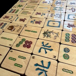 Puede incluir: Un conjunto de fichas de Mahjong de madera con varios s&iacute;mbolos y caracteres en verde, rojo y azul. Algunas fichas presentan dise&ntilde;os intrincados, mientras que otras muestran n&uacute;meros y palos. Las fichas est&aacute;n dispuestas sobre una superficie.
