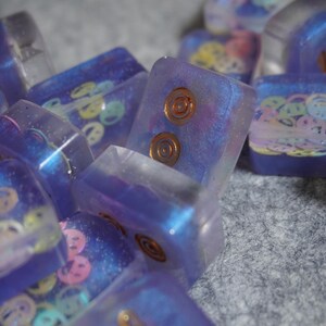 Pu&ograve; includere: Primo piano di diversi tasti rettangolari in resina traslucida. I tasti sono principalmente viola con disegni incorporati, tra cui faccine sorridenti e intarsi circolari color rame. I tasti hanno un effetto glitterato e iridescente.