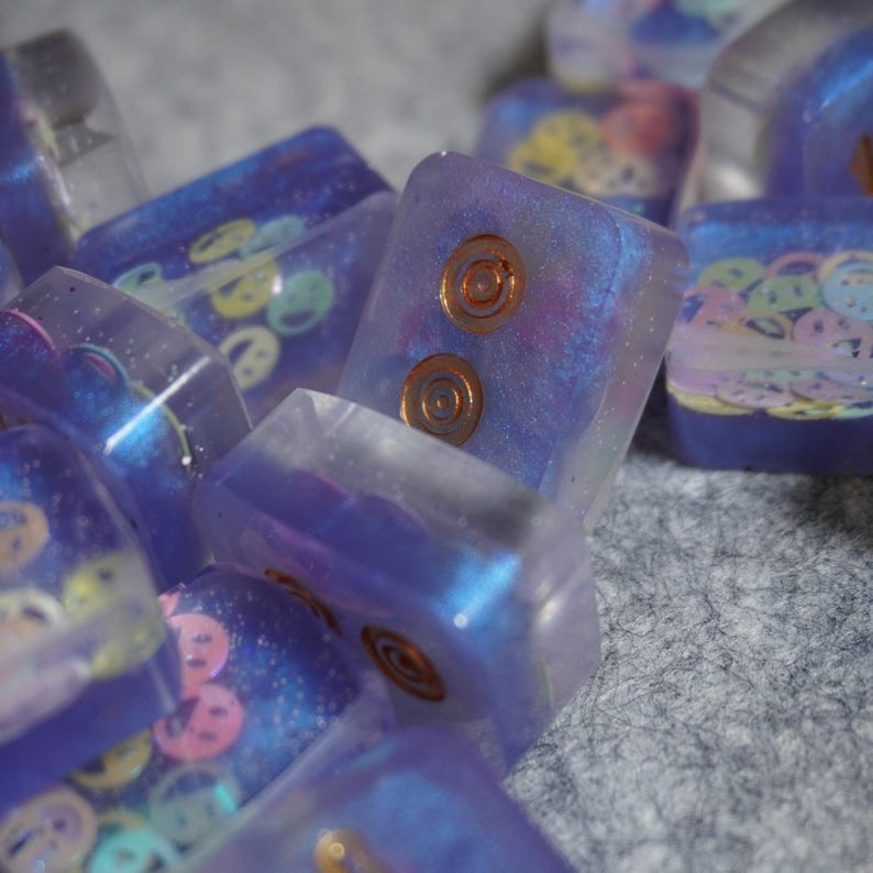 Pu&ograve; includere: Primo piano di diversi tasti rettangolari in resina traslucida. I tasti sono principalmente viola con disegni incorporati, tra cui faccine sorridenti e intarsi circolari color rame. I tasti hanno un effetto glitterato e iridescente.