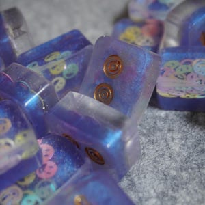 May include: Close-up of several rectangular, translucent resin keycaps. The keycaps are primarily purple with embedded designs, including smiley faces and copper-colored circular accents. Some keycaps have a shimmering, iridescent quality.