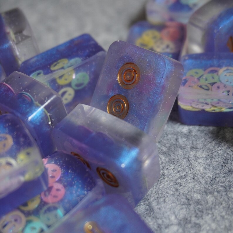 May include: Close-up of several rectangular, translucent resin keycaps. The keycaps are primarily purple with embedded designs, including smiley faces and copper-colored circular accents. Some keycaps have a shimmering, iridescent quality.