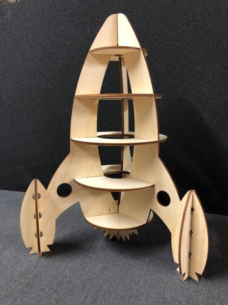 May include: A wooden rocket ship shelf unit with five shelves. The rocket ship is made of natural wood and has a light brown color. The shelves are all circular and are evenly spaced apart. The rocket ship has a pointed nose and two fins on the back.