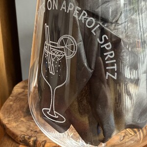 May include: A clear glass etched with the words "ITS ON APEROL SPRITZ". The glass has a detailed illustration of an Aperol Spritz cocktail with a straw and a lemon slice. The glass is partially filled with a dark material, possibly a napkin.