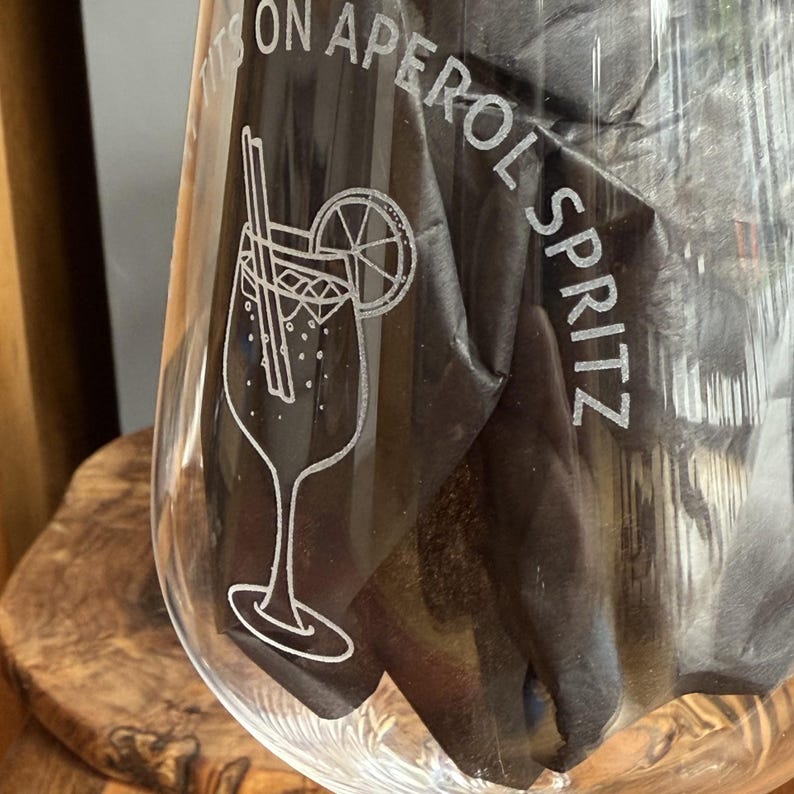 May include: A clear glass etched with the words "ITS ON APEROL SPRITZ". The glass has a detailed illustration of an Aperol Spritz cocktail with a straw and a lemon slice. The glass is partially filled with a dark material, possibly a napkin.