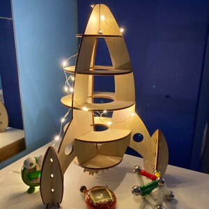 May include: A wooden rocket ship shelf with lights strung around it. The shelf has four levels and is made of light wood. The rocket ship is sitting on a white surface.