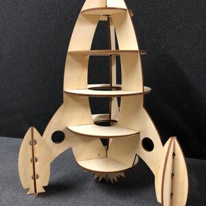 May include: A wooden rocket ship shelf unit with five shelves. The rocket ship is made of natural wood and has a light brown color. The shelves are all the same size and are evenly spaced. The rocket ship has a pointed nose and two fins on the back.