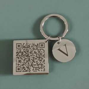 May include: Silver keyring with a square tag featuring a black and white QR code and a round tag with the letter "V" engraved on it.