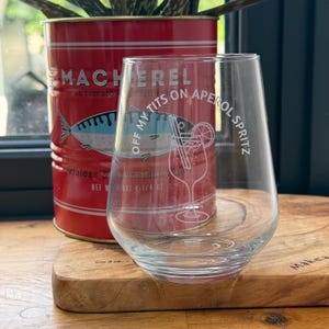 May include: A clear stemless wine glass with the text "OFF MY TITS ON APEROL SPRITZ" and a cocktail illustration. A red tin of mackerel in tomato sauce is behind the glass. The tin has a fish illustration and text.