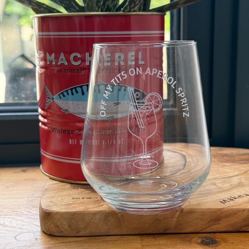 May include: A clear stemless wine glass with the text "OFF MY TITS ON APEROL SPRITZ" and a cocktail illustration. A red tin of mackerel in tomato sauce is behind the glass. The tin has a fish illustration and text.