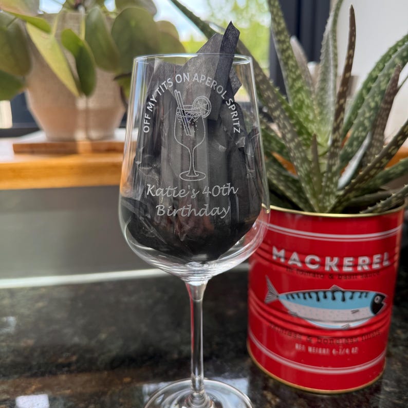 May include: A clear wine glass with etched text reading "OFF MY TITS ON APEROL SPRITZ" and "Katie's 40th Birthday". The glass is filled with black tissue paper. A red can with a mackerel label is visible in the background.