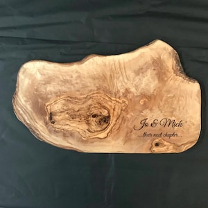 May include: A large, natural wood serving board with a unique, irregular shape. The board has a natural, rustic finish and is engraved with the text "Jo & Mick... their next chapter..."