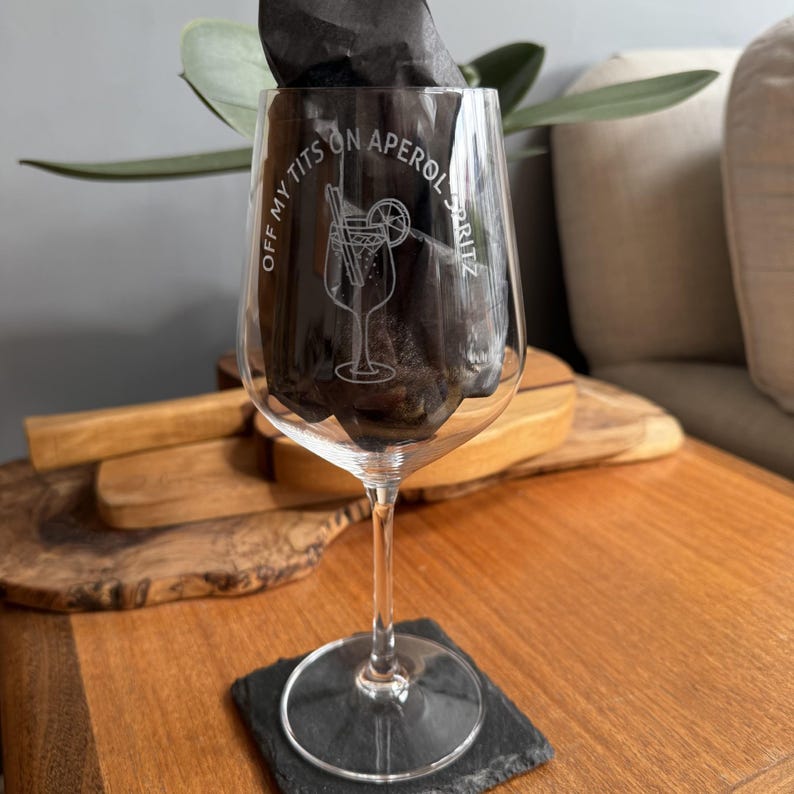 May include: A clear wine glass with etched text and a cocktail design. The text reads "OFF MY TITS ON APEROL SPRITZ". The glass is filled with dark contents and stands on a black square coaster. The glass has a long stem.