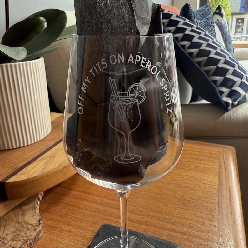 May include: A clear wine glass with a stem, etched with a cocktail design and the phrase "OFF MY TITS ON APEROL SPRITZ". The glass features a detailed illustration of a cocktail with a lemon slice. The glass is on a wooden table.