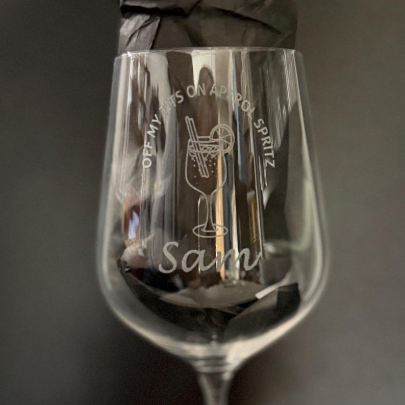 May include: A clear wine glass with etched text and a cocktail illustration. The text reads "OFF MY RITZ ON APEROL SPRITZ" and "Sam". The cocktail design includes a straw and a lemon slice.