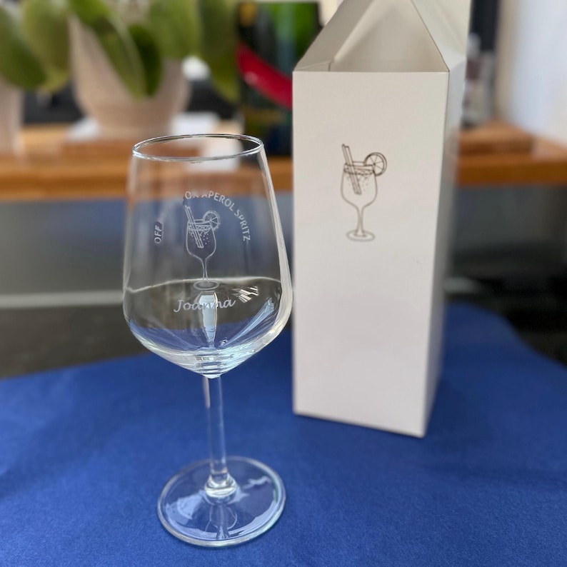 May include: A clear wine glass with a stem, featuring an etched cocktail design and the text "ON APEROL SPRITZ" and "OFF". A white box with a matching cocktail design is behind the glass. The name "Johanna" is etched on the glass.