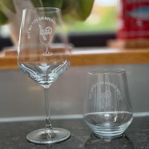 May include: Two clear wine glasses, one stemmed and one stemless, with the text "OFF MY TITS ON APERO" and a cocktail illustration. The glasses are on a dark countertop with a blurred background.