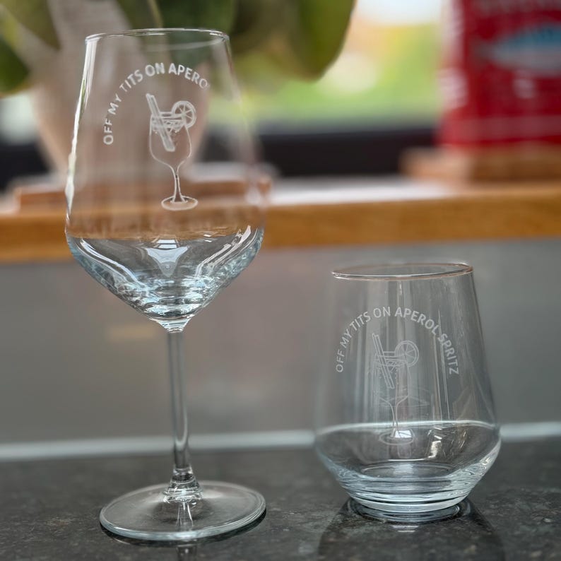 May include: Two clear wine glasses, one stemmed and one stemless, with the text "OFF MY TITS ON APERO" and a cocktail illustration. The glasses are on a dark countertop with a blurred background.