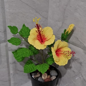 May include: A crocheted yellow hibiscus flower plant with red centres in a black pot with small stones. The plant is on a grey tiled surface.